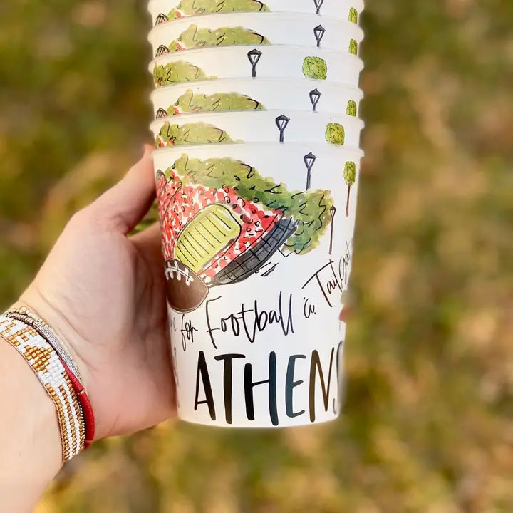 A hand holding a stack of white paper cups printed with a colorful illustration of a football field, football, trees, and the text 'for Football in Town ATHENS'. The background is a blurred outdoor scene.