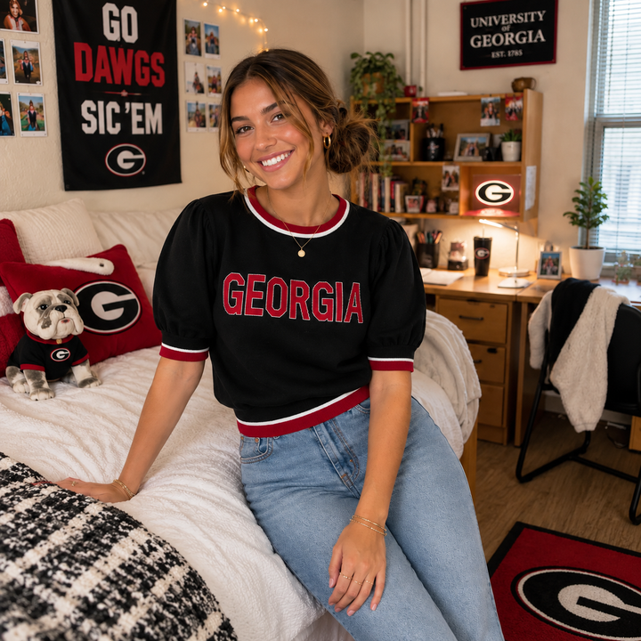 A woman sitting on a bed in a decorated bedroom wearing a black short-sleeve Georgia jersey shirt with red and white lettering, paired with light blue jeans. The room features University of Georgia themed decor, including a Bulldogs pillow, a dog toy, and a Georgia banner.