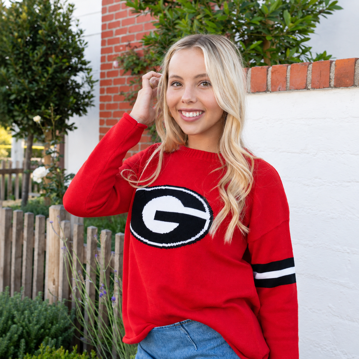 A smiling blonde woman wearing a bright red knit sweater with a large black and white Georgia 'G' logo on the chest and black-and-white striped sleeves, standing outdoors against a white wall and brick column.