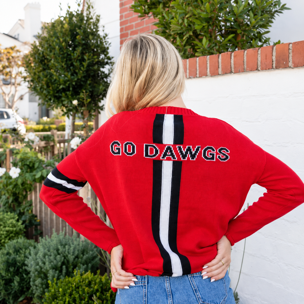 A woman viewed from behind wearing a red crewneck sweater with a vertical black and white stripe down the center and 'GO DAWGS' embroidered across the shoulders. She is standing outdoors in front of a white brick wall and green bushes.
