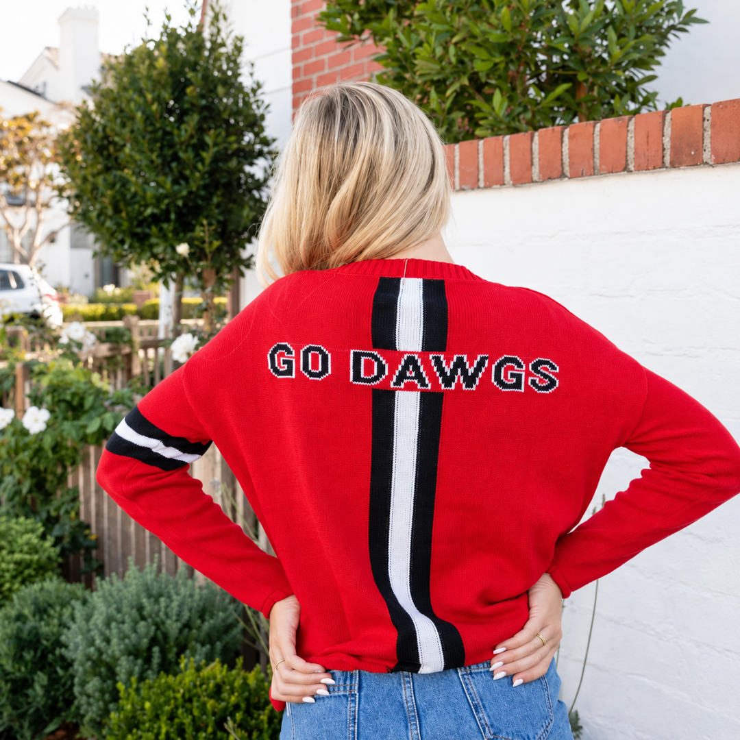 A woman viewed from behind wearing a red crewneck sweater with a vertical black and white stripe down the center and 'GO DAWGS' embroidered across the shoulders. She is standing outdoors in front of a white brick wall and green bushes.