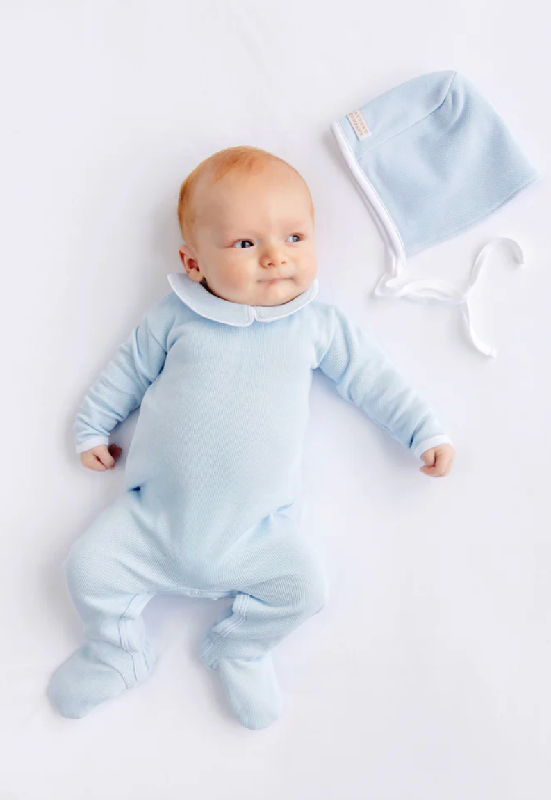 A baby lying on a white background wearing a light blue footed sleeper and a matching bonnet with white ties.
