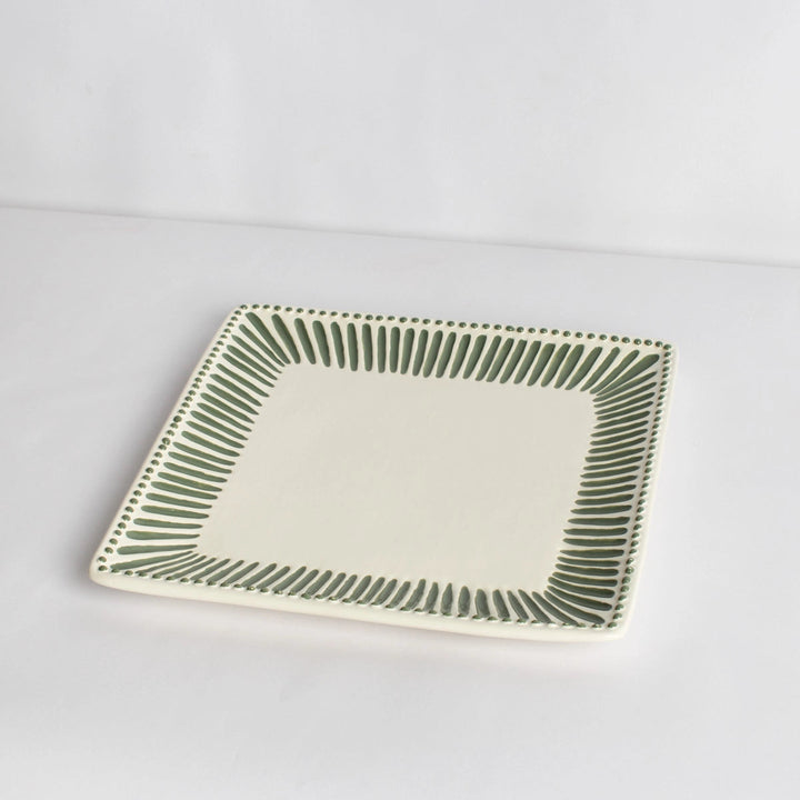 A square ceramic plate with a cream-colored center and a green hand-painted dotted border, photographed on a plain white background.